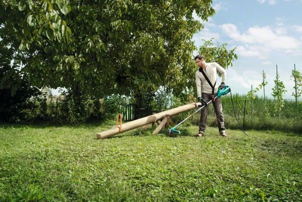 Muškarac s Bosch AFS 23-37 trimerom za travu obrezivanje trave u vrtu pored stabla.