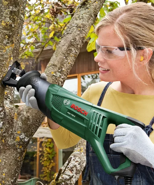Žena s zaštitnim naočalama i rukavicama koristi Bosch PSA 700 E sabljastu pilu na drvetu.