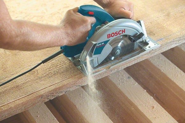 Ruka drži BOSCH GKS 65 ručnu kružnu pilu dok reže drvene grede, ispuštajući piljevinu.