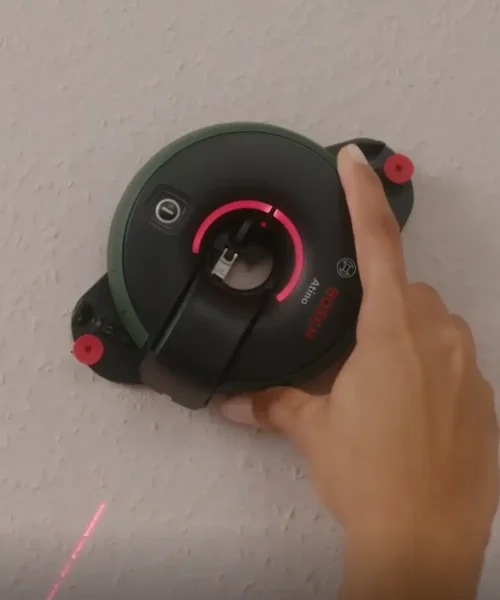 Ruka drži BOSCH laserski nivelir Atino 2 s metrom, prikazan na zidu za niveliranje.